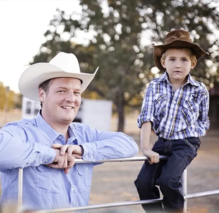 A man in a white cowboy hat and a boy in a brown hat at a metal ranch gate under an oak tree